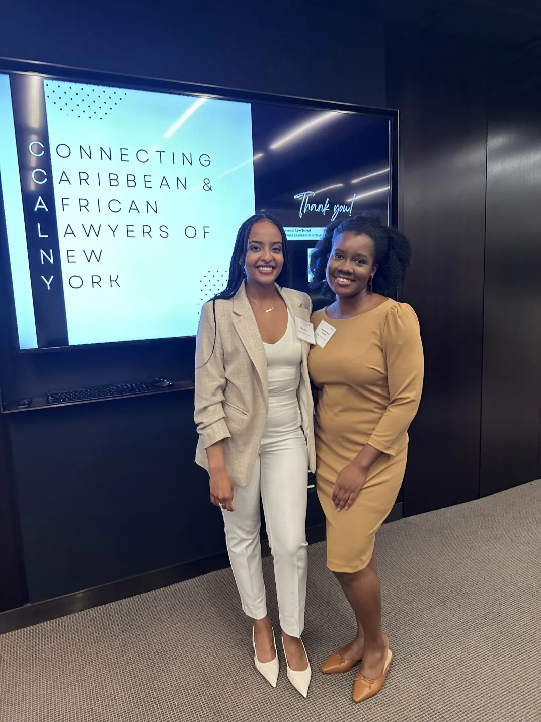 Tobin Kassa is wearing a white top with a beige blazer, white pants and white heels. She is standing on the left, with Dawchelle Hamilton to the right. Dawchelle is wearing a yellow-brown colored dress with brown flats. They are both standing in front of a TV with there event logo, and both are smiling for their photo.